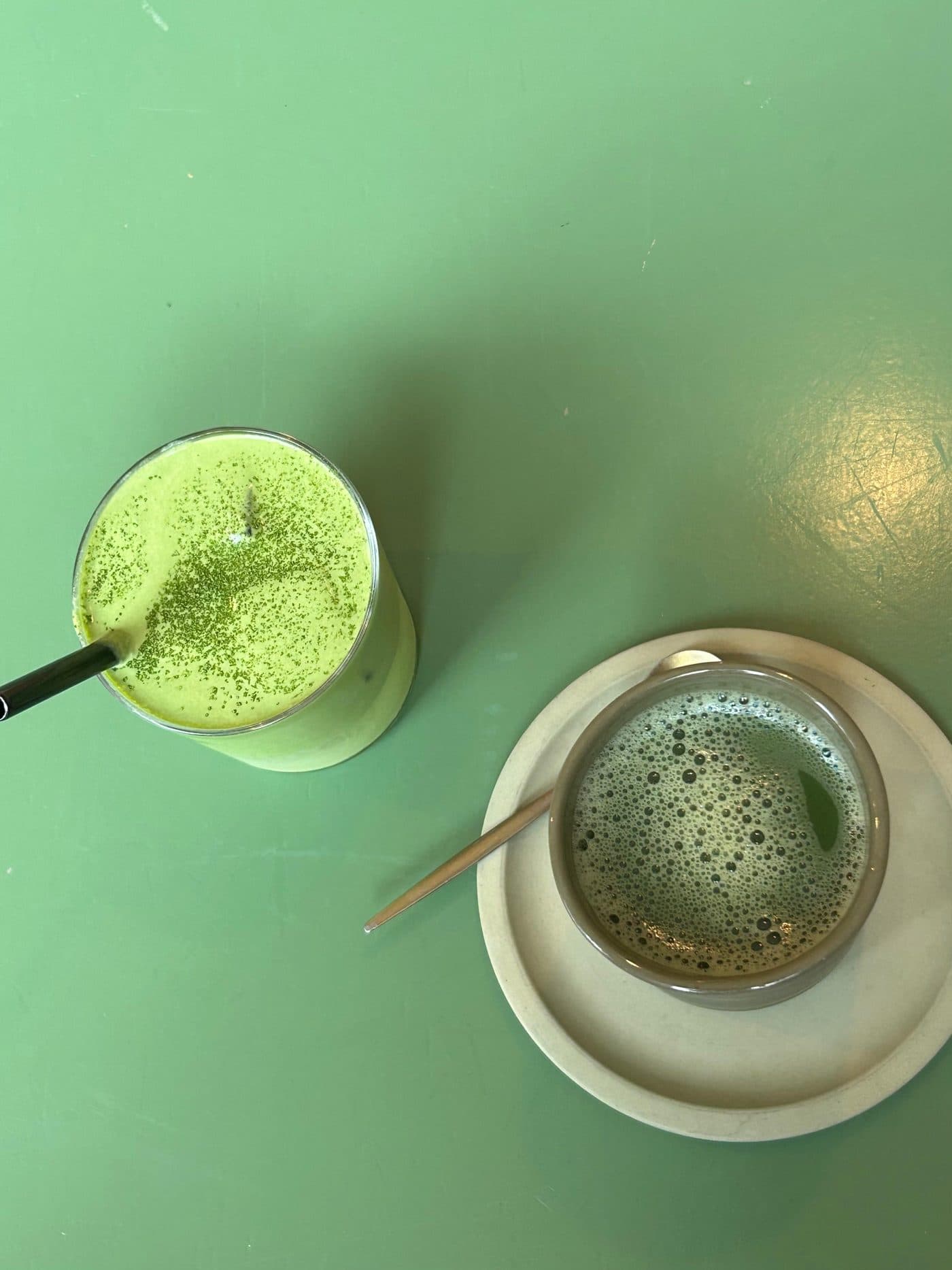 Un matcha latte en vaso y una taza de matcha sobre una mesa verde
